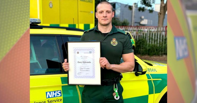 Gary Edwards in uniform stood in front of an ambulance car holding a certificate