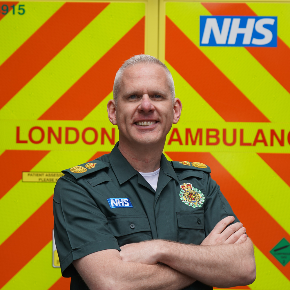 A photo of Craig Harman in his green ambulance uniform, in front of an emergency ambulance.