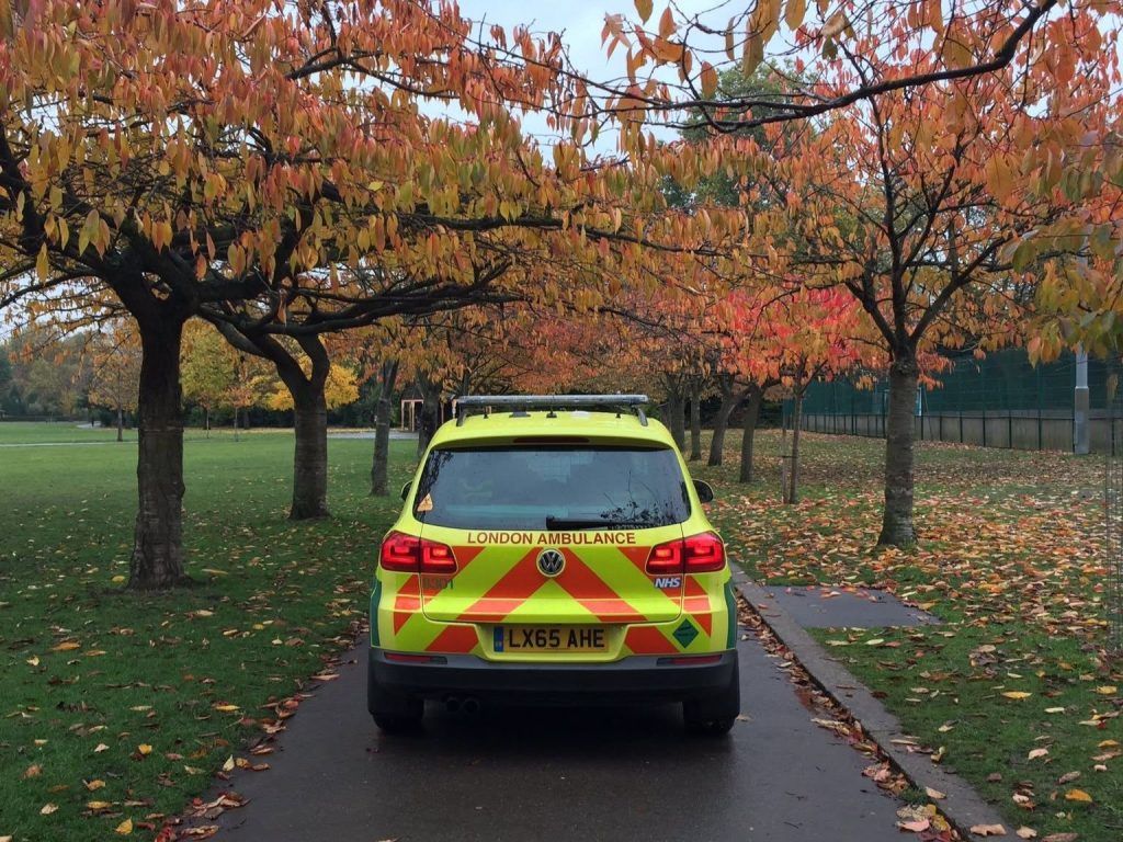 A London Ambulance Service response car on a park road surrounded by trees with brown autumn leaves.
